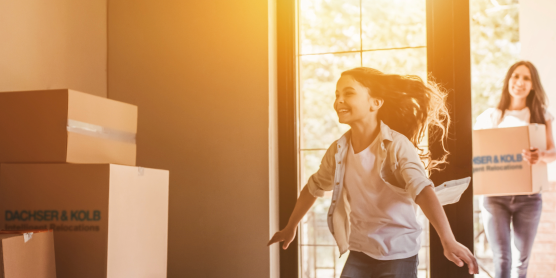 Fröhliche Familie beim Privatumzug – Kind läuft lachend ins neue Zuhause, Eltern tragen Umzugskartons ins Haus.