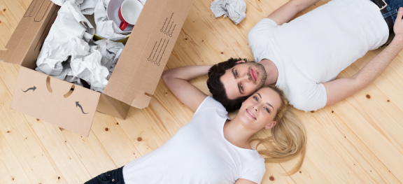 Glückliches Paar liegt entspannt neben ausgepacktem Karton. Prioritäten beim Umzug: Wichtiges zuerst auspacken, Zeit nehmen.
