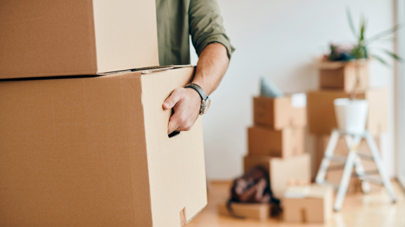 Person trägt Umzugskarton in Wohnung – Umzugskartons im Hintergrund, Symbol für Umzug und Organisation