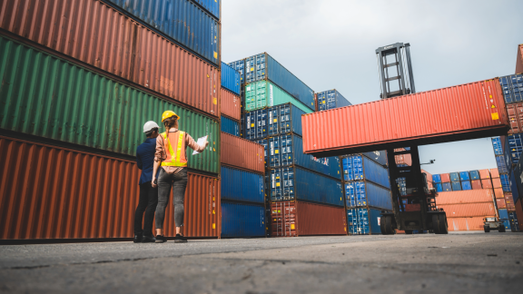 Zwei Hafenarbeiter stehen vor gestapelten bunten Containern. Ein Stapler bewegt einen FCL-Container.
